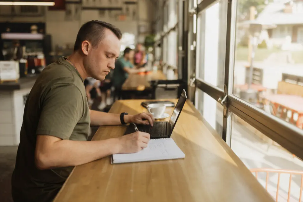 male adult learner taking notes in a notebook while watching a lecture on a laptop
